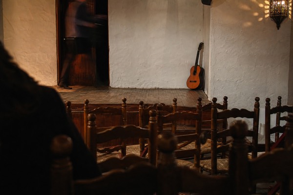 Bare wooden tablao stage with empty chairs and a guitar resting against a whitewashed wall in Santa Cruz, Seville