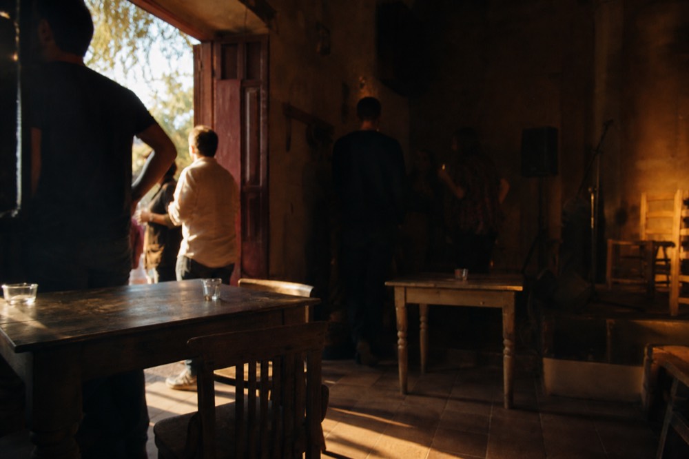Rustic Seville bar interior with mismatched wooden tables, a bare stage corner and people standing with drinks in low light