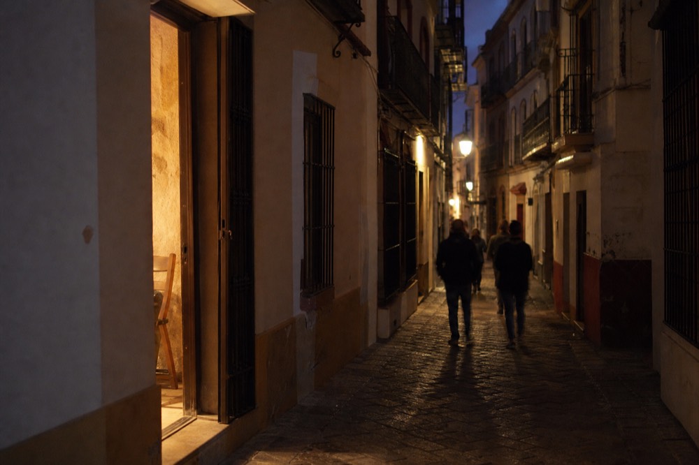 Narrow Seville street at night with warm light spilling from an open doorway onto the cobbles, no signage or lettering visible