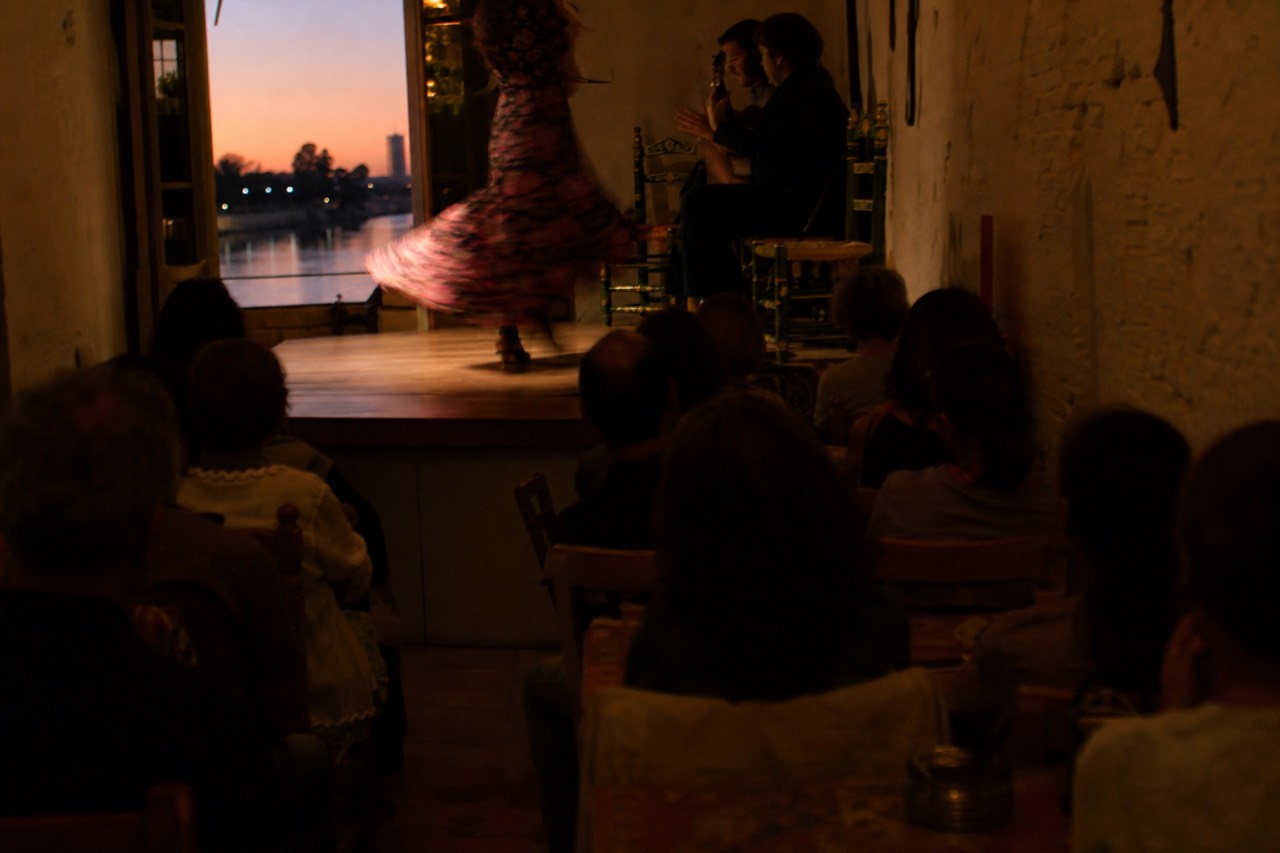 Narrow riverside tablao room with a low wooden stage and close-packed chairs, a dancer's skirt caught mid-turn in blur
