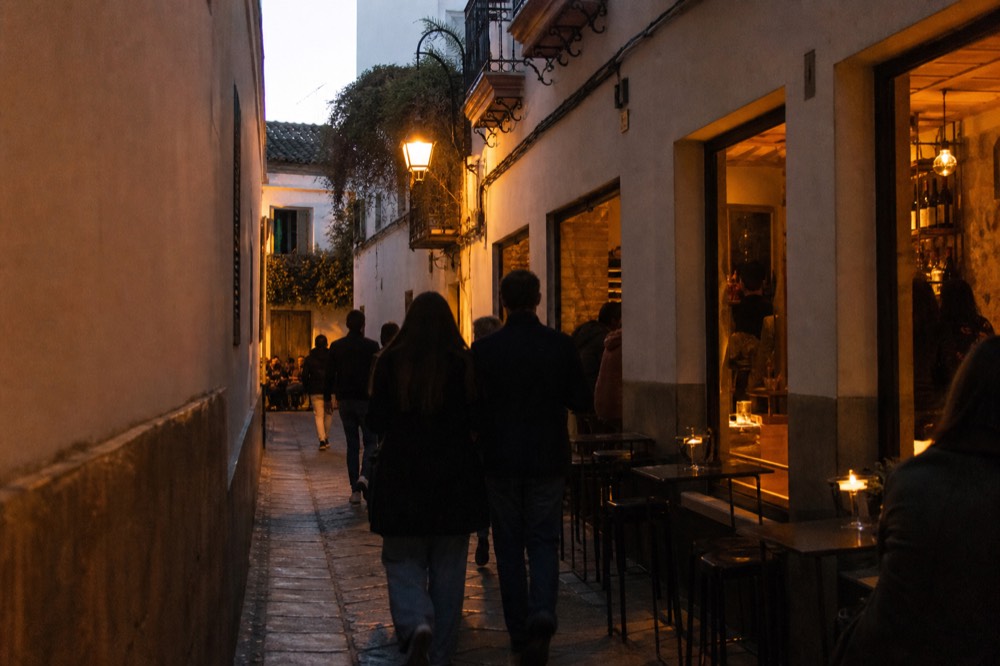 A narrow Seville street at dusk with warm lit bar windows, small tables on the pavement and a few figures walking towards the sound of guitar