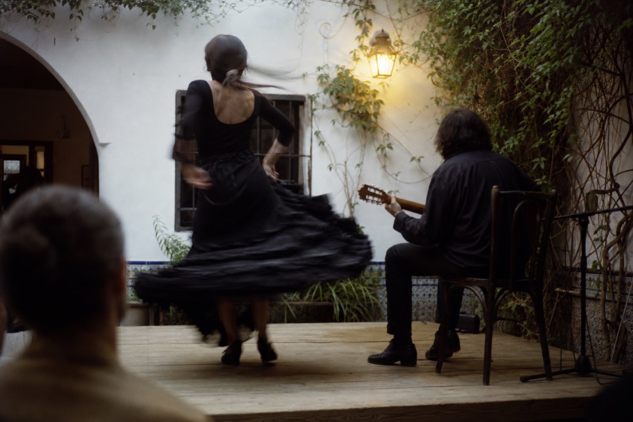 A flamenco dancer photographed from behind mid-turn on a small wooden stage, skirt caught in motion blur, a seated guitarist behind her under warm lamplight