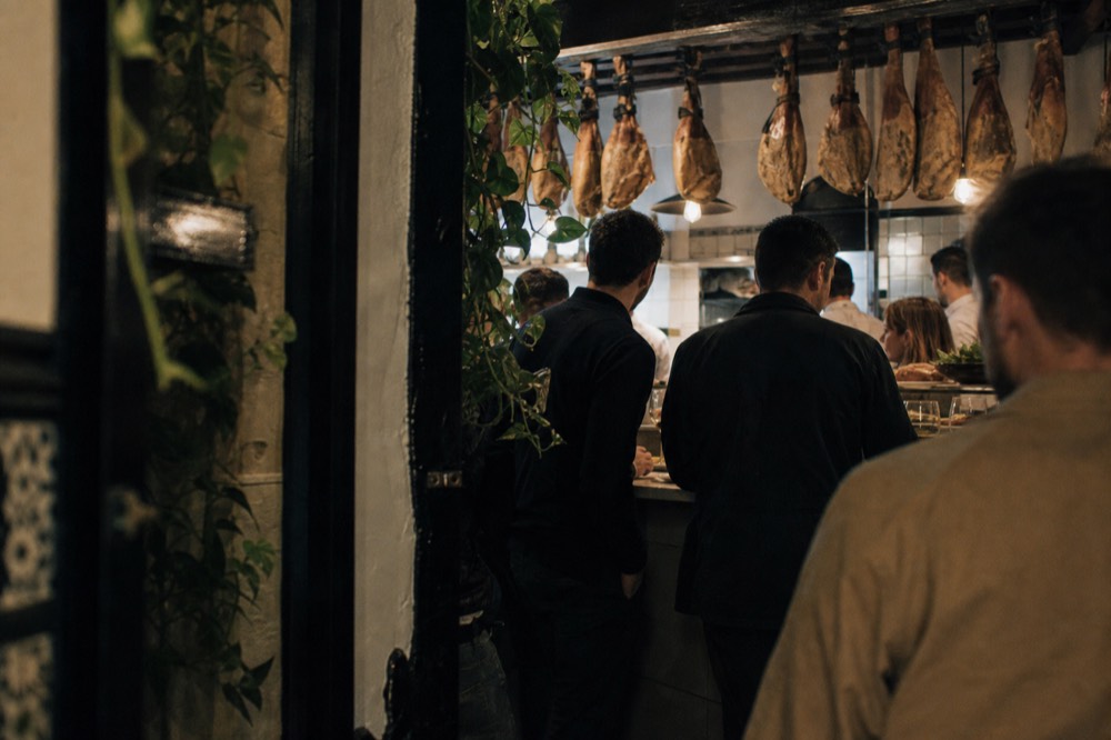 A busy late-night tapas bar counter seen from the doorway, hams hanging above, customers standing shoulder to shoulder with small plates