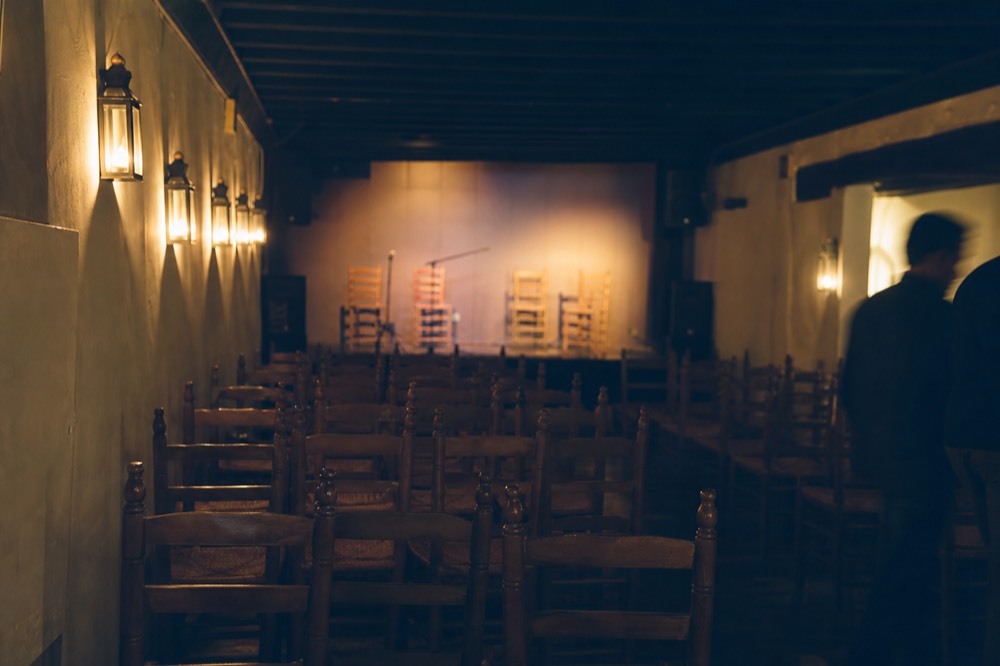 Small tablao room with wooden chairs in tight rows facing a low stage, empty before the doors open, warm lamps along the wall