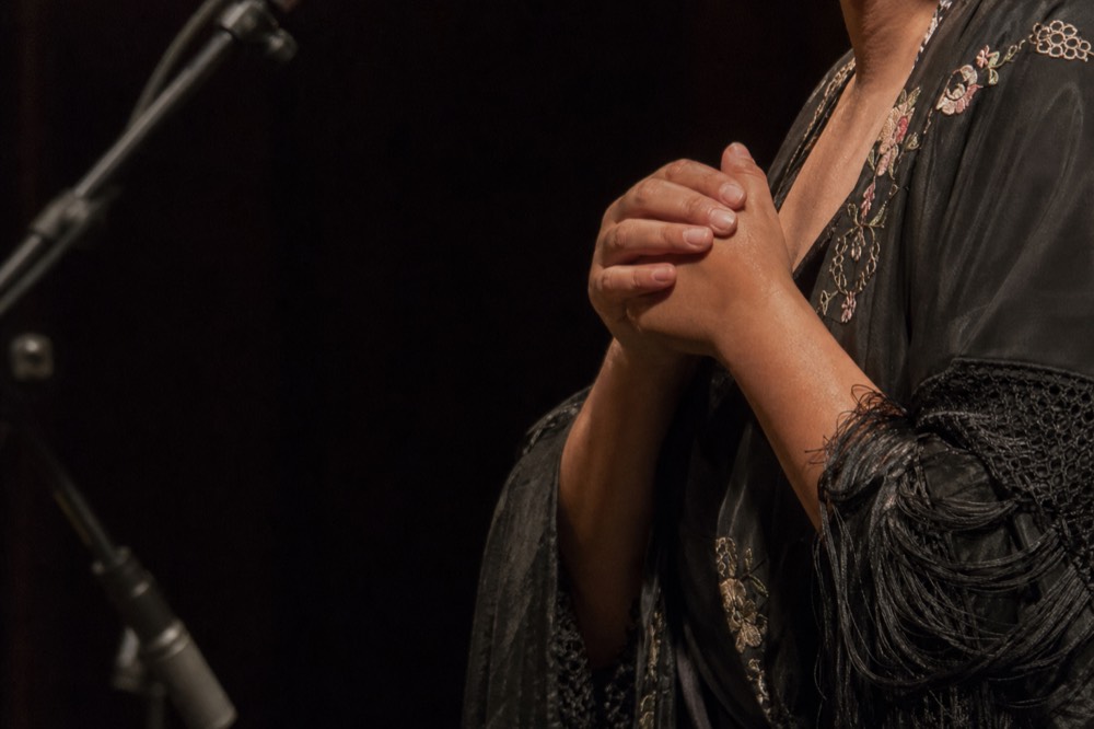 Close crop of a singer's hands clasped at chest height under stage light, face out of frame, dark background