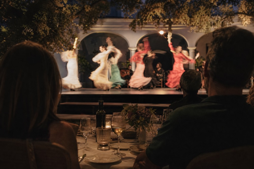 Wide theater-scale flamenco stage seen from a rear dinner table, a line of dancers in motion blur under warm spotlights