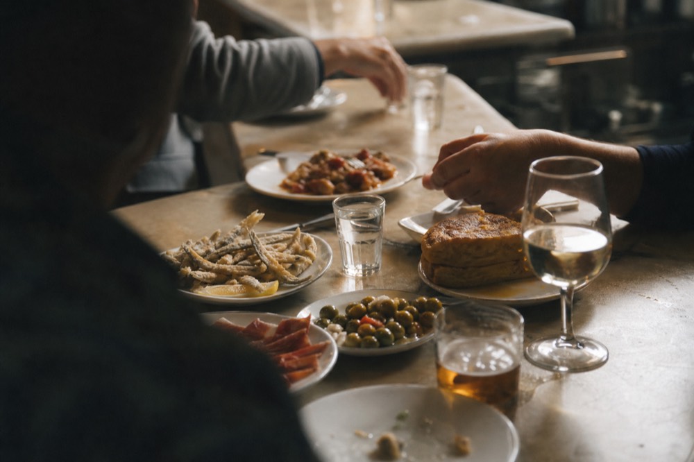 Tapas plates and small glasses crowded on a marble bar top in Seville, hands reaching in from the edges of the frame, no faces