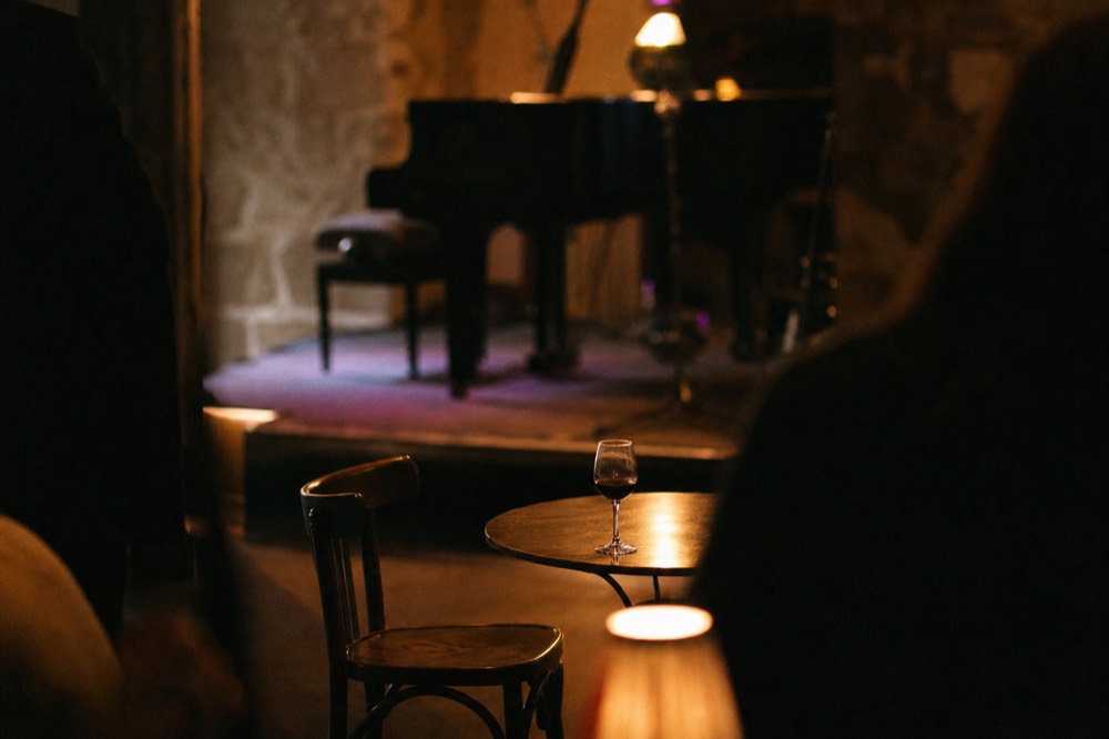 Small round table with a single glass of wine beside an empty chair, blurred stage lighting behind, dark intimate room