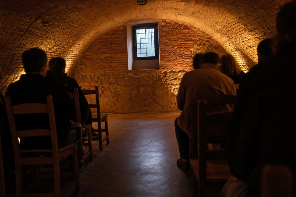 Two rows of simple wooden chairs facing a bare floor inside a low brick vault, warm uplighting on the curved ceiling