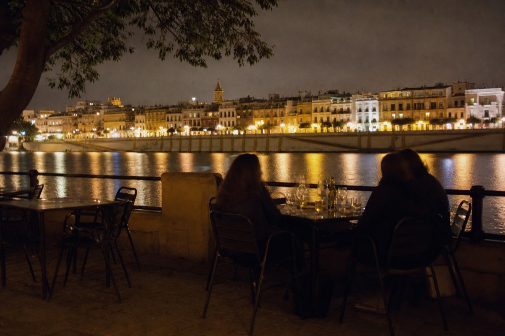 Riverside terrace tables on Calle Betis in Triana at night, the Guadalquivir reflecting the lit facades of the old town opposite
