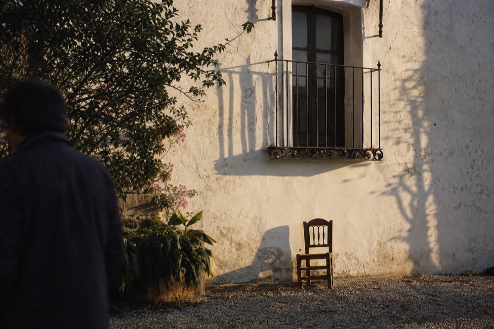 Weathered whitewashed wall of an old Andalusian courtyard with an iron balcony and a single wooden chair below it in late afternoon light
