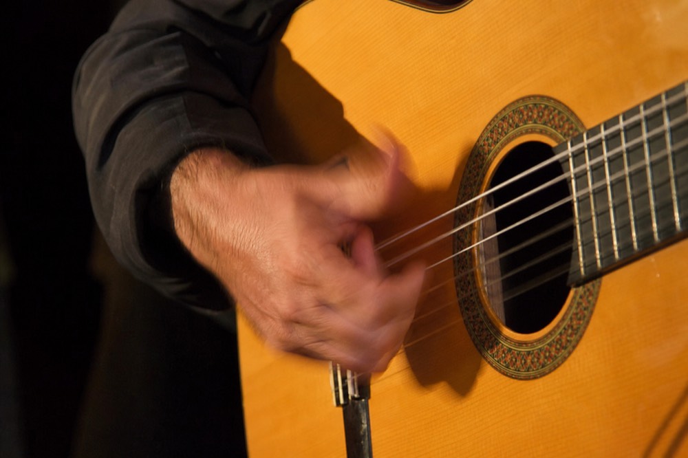 Close crop of a flamenco guitarist's right hand mid-rasgueado over the soundhole, strings blurred, nails catching the stage light